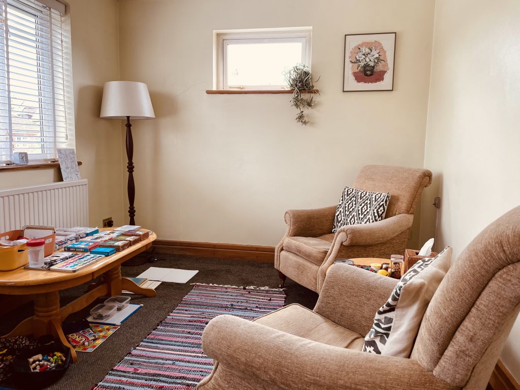 Cosy therapy room featuring comfortable seating and a table with sensory, art, and play materials.